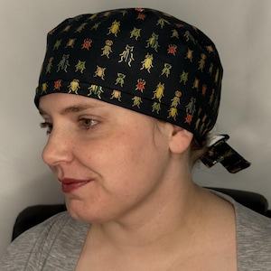 May include: A black scrub cap featuring a repeating pattern of small, colorful beetles in shades of yellow, green, orange, and blue. The cap has a wide, folded front band and ties at the back to secure the fit, covering the top and back of the head.