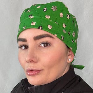 May include: A green scrub cap featuring a repeating pattern of small illustrations including frogs, cats, diamonds, and flowers. The fabric head covering has a folded front band and ties at the back to secure the fit, providing full coverage for the head.