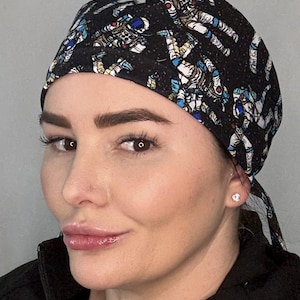 May include: A scrub cap featuring a black background with a repeating pattern of astronauts in white suits with blue accents, floating amidst a field of small white stars. The cap has a fitted crown and tie-back closure for a secure fit.
