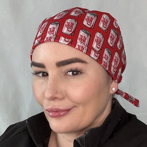 May include: A red scrub cap featuring an all-over repeating pattern of silver and white Diet Coke soda cans. The fabric head covering has a fitted crown and includes two fabric ties at the back to secure the fit, displaying the brand logo clearly.