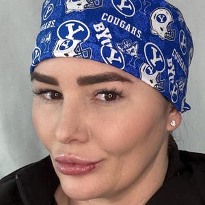 Peut inclure: Une charlotte de bloc opératoire bleue avec un motif répétitif de logos blancs Brigham Young University Cougars, de casques de football américain et du texte BYU et COUGARS. Le tissu présente un fond bleu texturé avec des éléments graphiques blancs.