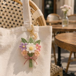 May include: A cream-colored canvas tote bag featuring a hanging keychain ornament. The ornament is a handcrafted yarn bouquet with green leaves, a white daisy, a purple flower, and two pink flowers, tied with a pink yarn bow and a gold butterfly charm.