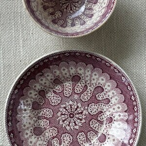 May include: Two ceramic bowls with a white background and a burgundy floral pattern. The smaller bowl is on top of the larger bowl.