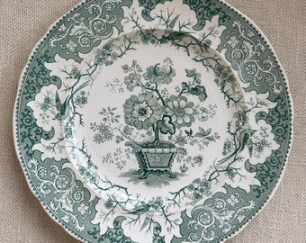 May include: A vintage ceramic plate with a green floral design. The plate features a central image of a potted plant with flowers and leaves, surrounded by a decorative border of swirling vines and birds.