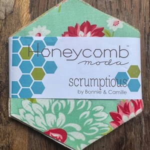 Puede incluir: Una pila de piezas de tela hexagonales con un fondo verde claro y patrones florales rojos y blancos. Una banda de papel blanca envuelve el centro, impresa con el texto Honeycomb moda scrumptious by Bonnie & Camille.