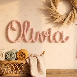 May include: A wall-mounted name sign spelling Olivia in soft pink knitted yarn script. Below, a woven rattan basket holds three rolled blankets in cream, sage green, and mustard yellow, with a beige fabric draped over the basket edge.