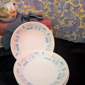 May include: Two white ceramic bowls featuring a repeating pattern of light blue and grey abstract geometric shapes and stylized furniture icons around the interior rim. The bowls have a smooth, rounded edge and a clean, white center basin.