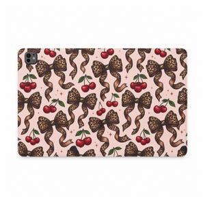 Peut inclure: Une coque de tablette rectangulaire présentant un motif répétitif sur un fond rose clair. Le design se compose de nœuds à imprimé léopard marron et de paires de cerises rouge vif avec des tiges vertes, agrémentées de petites étoiles.