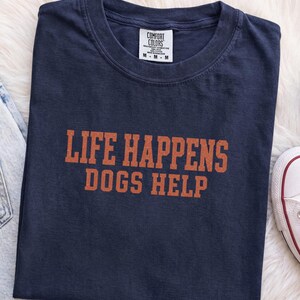 May include: A navy blue short-sleeve t-shirt with a crew neckline. The shirt features the orange text "LIFE HAPPENS DOGS HELP" printed in a bold, block font across the chest. A white label inside the collar reads "COMFORT COLORS" with size M.