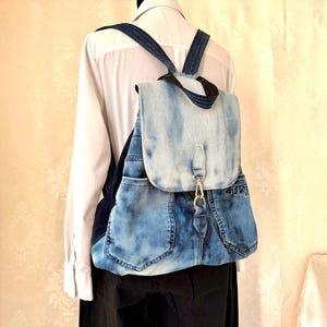 May include: A backpack constructed from repurposed blue denim fabric featuring a light-wash flap with a gold-toned metal lobster clasp closure, two front pockets, and dark blue denim shoulder straps, worn over a white button-down shirt.