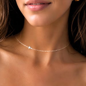 May include: A delicate gold-colored necklace featuring a small cross pendant. The cross is positioned slightly off-center on a fine chain. The necklace is designed to sit close to the neckline, creating a subtle and elegant look.