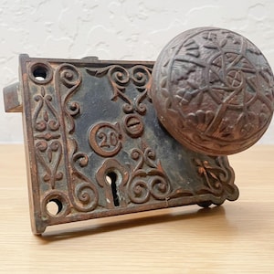 May include: Antique cast iron door lock set with a round, patterned doorknob. The lock plate is rectangular with ornate scrollwork and the number "20" embossed. The finish is a weathered brown with signs of age.