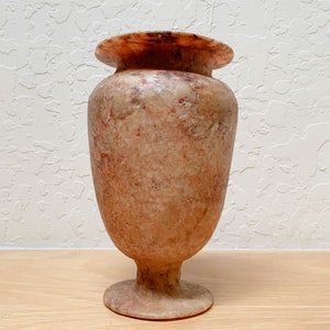 May include: A decorative vase with a wide, flared rim and a rounded body. The vase is made of a stone material with a mottled pattern of beige, brown, and reddish-pink hues. It has a pedestal base and a smooth, polished finish.