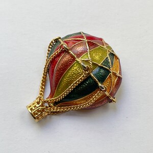 May include: A colorful hot air balloon brooch with a gold chain basket. The balloon is divided into sections of red, green, yellow, and orange. The brooch is made of metal and has a shiny finish.