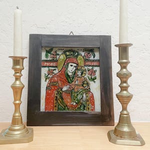 May include: A square wooden frame containing a religious icon painting of a figure holding a child, flanked by two tall brass candlesticks with white taper candles. The painting features red, green, and gold tones with text reading CMNOY and 1829.