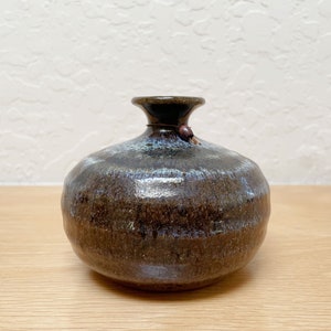 May include: A small, round ceramic vase with a dark brown base and a lighter, blue-tinged top. The vase has a narrow neck and a small, decorative bead attached. The glaze has a textured appearance, giving it a rustic feel.