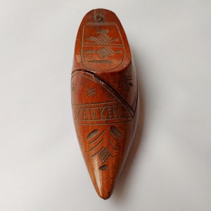 May include: A wooden shoe-shaped box with intricate carvings and the word "MARKEN" etched on the side. The box is a warm reddish-brown color and features decorative patterns on the top and toe. A small metal screw is visible near the top.