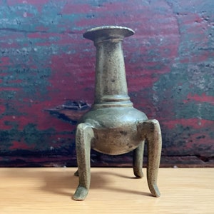 May include: A bronze-colored, antique candle holder with a flared top and three legs. The candle holder has a cylindrical body and a slightly weathered appearance, suggesting age and use. It stands on a wooden surface.