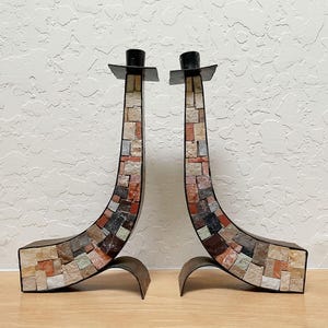 May include: Pair of decorative candle holders. Each holder has a mosaic-tiled, curved body in shades of red, brown, and gray, with a black top and base. The base has a curved, metal design.