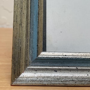 May include: Close-up of a picture frame corner. The frame has a distressed finish with shades of silver, blue, and gold. The frame's corner is a mitered joint, and the frame appears to be made of wood. The frame is empty.