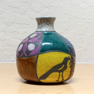 May include: A colorful ceramic vase with a patchwork design. The vase features a variety of colors, including brown, purple, green, and yellow. A black bird silhouette is on the yellow section. The vase has a cracked, textured finish.