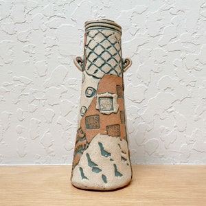 May include: A tall, tapered ceramic vase featuring a textured, off-white and terracotta-colored surface. The design includes a grid pattern at the top, two small side handles, and several square stamped impressions with intricate characters.