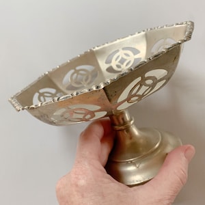 May include: A silver-toned, footed dish with an openwork design. The dish has a rectangular shape with rounded corners and a decorative edge. The base is a pedestal style. The dish is held by a hand.