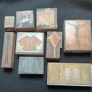 May include: A collection of vintage printing blocks, featuring various designs. The blocks are made of wood and metal, with images of a lamp post, clothing, and architectural elements. The blocks are in shades of brown and copper, with a dark background.