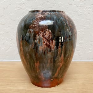 May include: A glazed ceramic vase with a rounded body and a wide opening. The vase features a blend of colors, including dark brown, black, blue, and reddish-brown hues. The glossy finish enhances the visual appeal of the decorative piece.