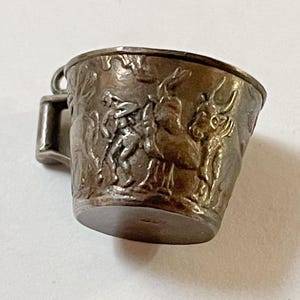 May include: A small, metallic, bronze-toned charm shaped like a miniature cup. The exterior surface features a detailed, raised relief design depicting figures and animals. A small loop is attached to the side, allowing it to be worn as a pendant.