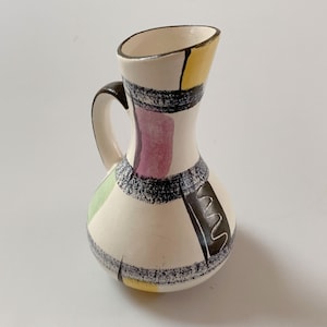 May include: A small white ceramic pitcher with a geometric design in black, yellow, green, and purple. The pitcher has a handle and a rounded body.