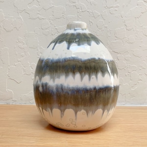 May include: A ceramic vase with a rounded body and a narrow neck. The vase features horizontal bands of white, blue, and olive green glaze. The base and neck are white, and the vase sits on a wooden surface.