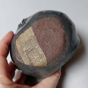 May include: A handcrafted ceramic bowl with a unique, organic shape. The bowl features a dark gray exterior with a glossy finish. The interior has a textured surface with a color block design of brown, tan, and gray. The bowl is held by a hand.