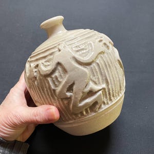 May include: A beige ceramic vase with a rounded body and a narrow neck. The vase features a relief design of stylized figures and vertical lines. The vase is held in a hand against a dark background.