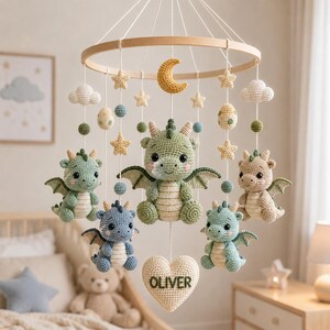 May include: A nursery mobile featuring five crocheted dragons in green, blue, and beige, suspended from a circular wooden frame. The mobile includes crocheted stars, clouds, a crescent moon, and a heart-shaped piece with the name OLIVER.