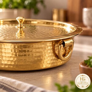 May include: A round, hammered brass serving dish with a matching lid and a central knob handle. The dish features two side-mounted ring handles for lifting. The surface has a reflective, metallic gold finish with a textured, dimpled pattern.