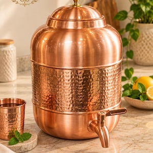 May include: A large copper water dispenser with a hammered texture band around the centre, a matching lid with a gold-toned knob, and a functional spigot. A smaller matching hammered copper cup sits to the left of the main vessel.