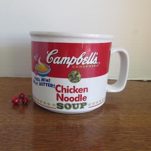 May include: A white ceramic mug with a red band featuring the Campbell's Condensed logo and Chicken Noodle Soup text. The mug has a handle and the words "Feel M'm! M'm! Better!" are printed on the side. A few red berries are on the wooden surface.