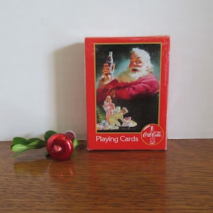 May include: A red box of Coca-Cola playing cards featuring a vintage illustration of Santa Claus holding a bottle of Coca-Cola. The box has the text "Playing Cards" and the Coca-Cola logo.