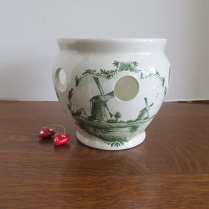 May include: White ceramic planter with a green windmill design. The planter has three circular openings and a flared base. Two red mushroom ornaments are in the foreground. The design features windmills, trees, and a landscape scene.