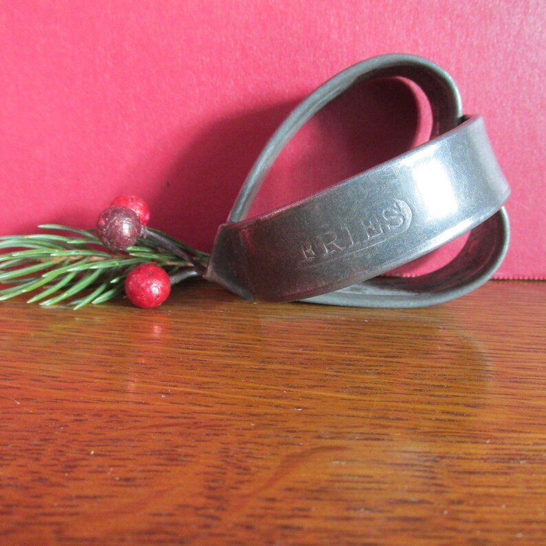 May include: A black leather belt buckle with the word "BRIMS" embossed on it. The buckle is in the shape of a heart and is lying on a wooden surface.