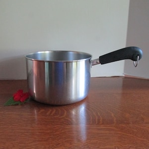 May include: A stainless steel saucepan with a polished silver finish and a black handle. The handle features a small metal loop at the end for hanging. The pot has a cylindrical shape with a flat base and straight sides, reflecting light on its surface.
