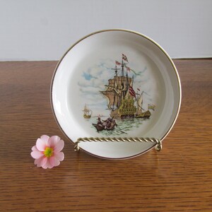 May include: A white ceramic coaster with a gold rim features a vintage illustration of a sailing ship with brown sails and a yellow hull. The ship is surrounded by blue water and a light blue sky.