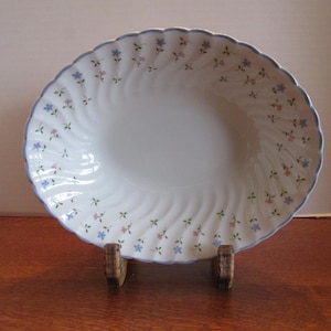 May include: White ceramic serving bowl with a blue scalloped edge and a floral pattern of pink and blue flowers. The bowl has a spiral design and is displayed on a wooden stand.
