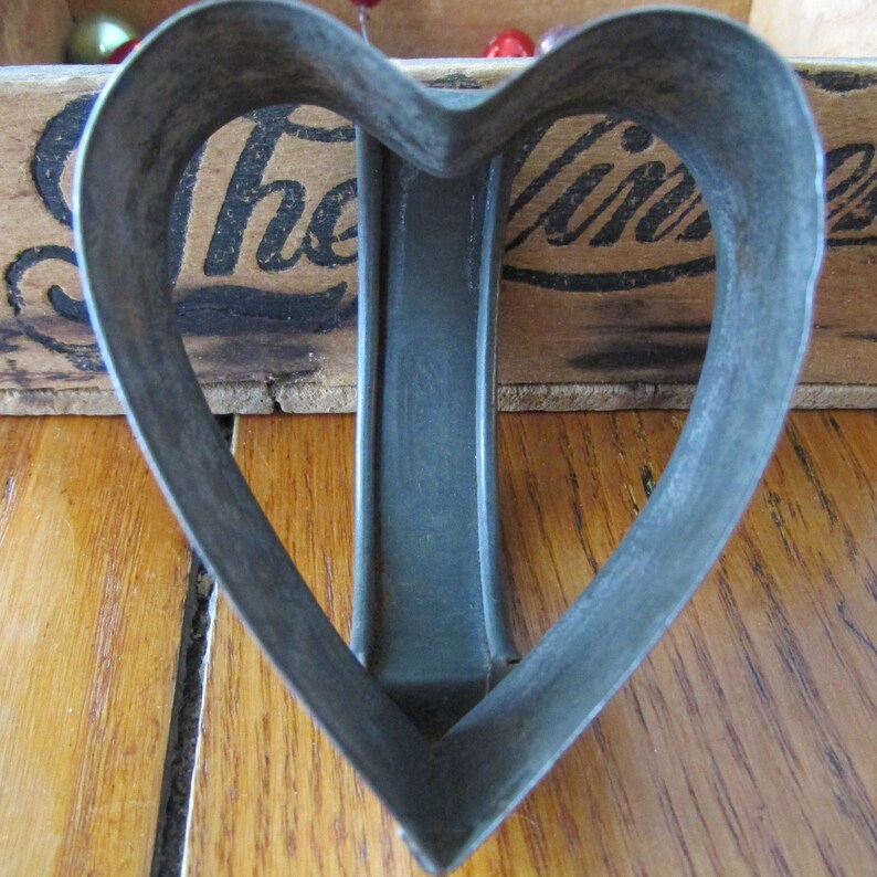 May include: A vintage metal heart-shaped cookie cutter. The cutter is made of a dark gray metal and has a smooth, slightly worn surface.