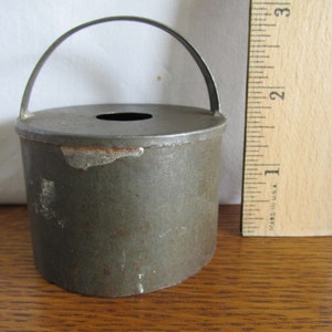May include: A vintage metal cookie cutter in the shape of a small pail with a handle. The cutter is gray and has a worn, rustic look.