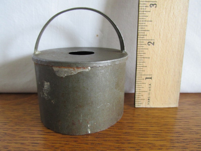 May include: A vintage metal cookie cutter in the shape of a small pail with a handle. The cutter is gray and has a worn, rustic look.