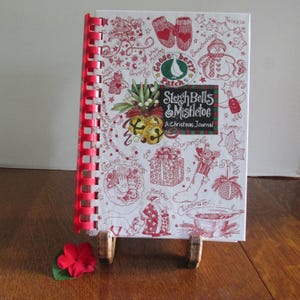 May include: A spiral-bound Christmas journal titled "Sleigh Bells & Mistletoe" with red and white illustrations. The cover features drawings of mittens, a snowman, bells, and a gift. The journal is propped up on a wooden stand, with a red flower in the foreground.