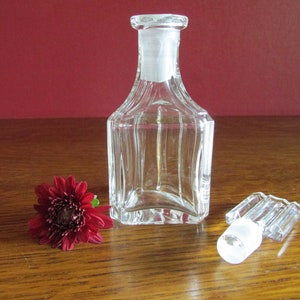 May include: A clear glass bottle with a square body and a textured surface. The bottle has a clear glass stopper with a ribbed design. The bottle and stopper are sitting on a wooden surface.