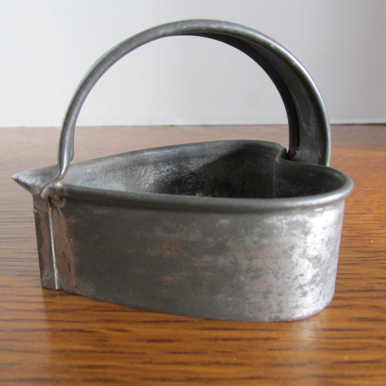 May include: A vintage metal heart-shaped cookie cutter with a handle. The cutter is made of silver-colored metal and has a worn, rustic look.